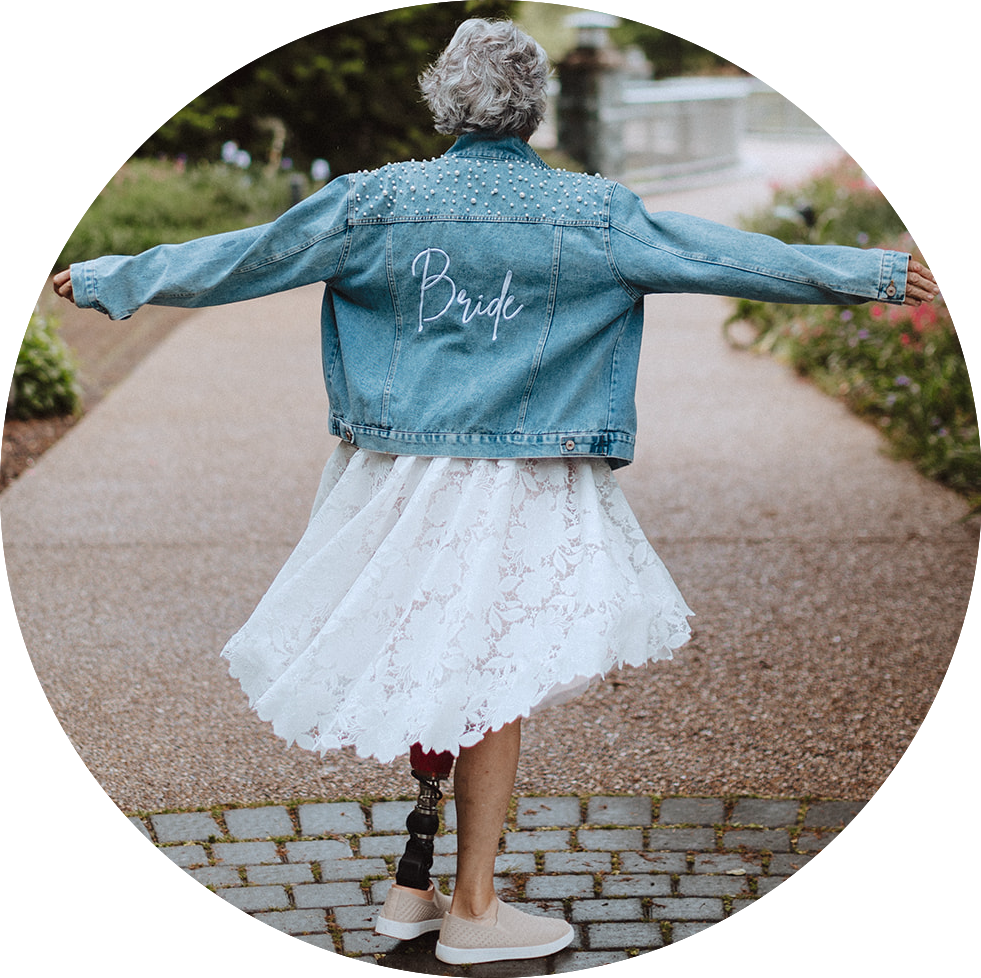 Sue, a white woman with short grey hair and a red prosthetic leg, spins in a jean jacket reading "Bride"