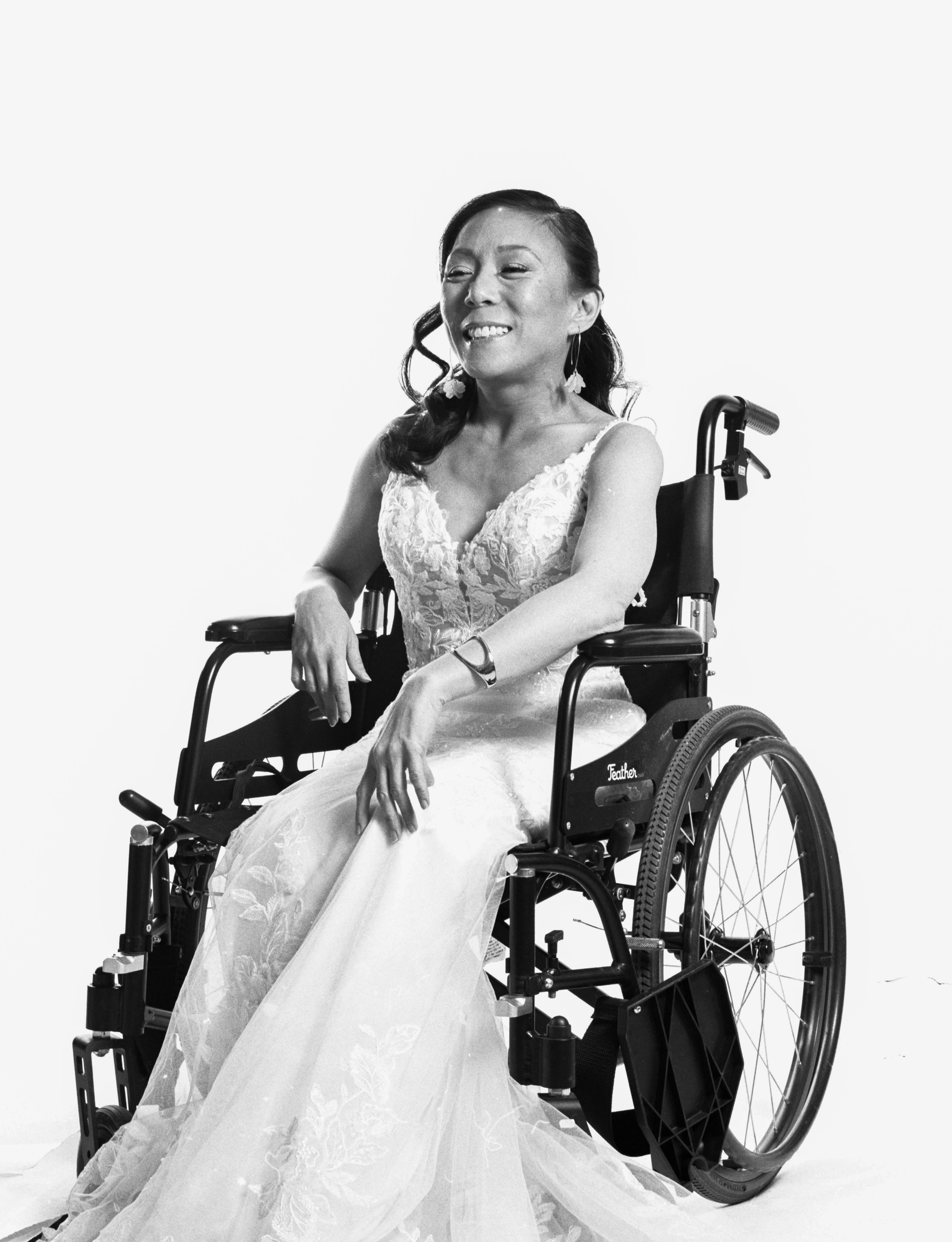 Dramatic black and white photo. Tamara, a short, dark-haired Asian woman wearing a fitted floral lace wedding dress, leans on the handles of her wheelchair smiling brightly at the camera