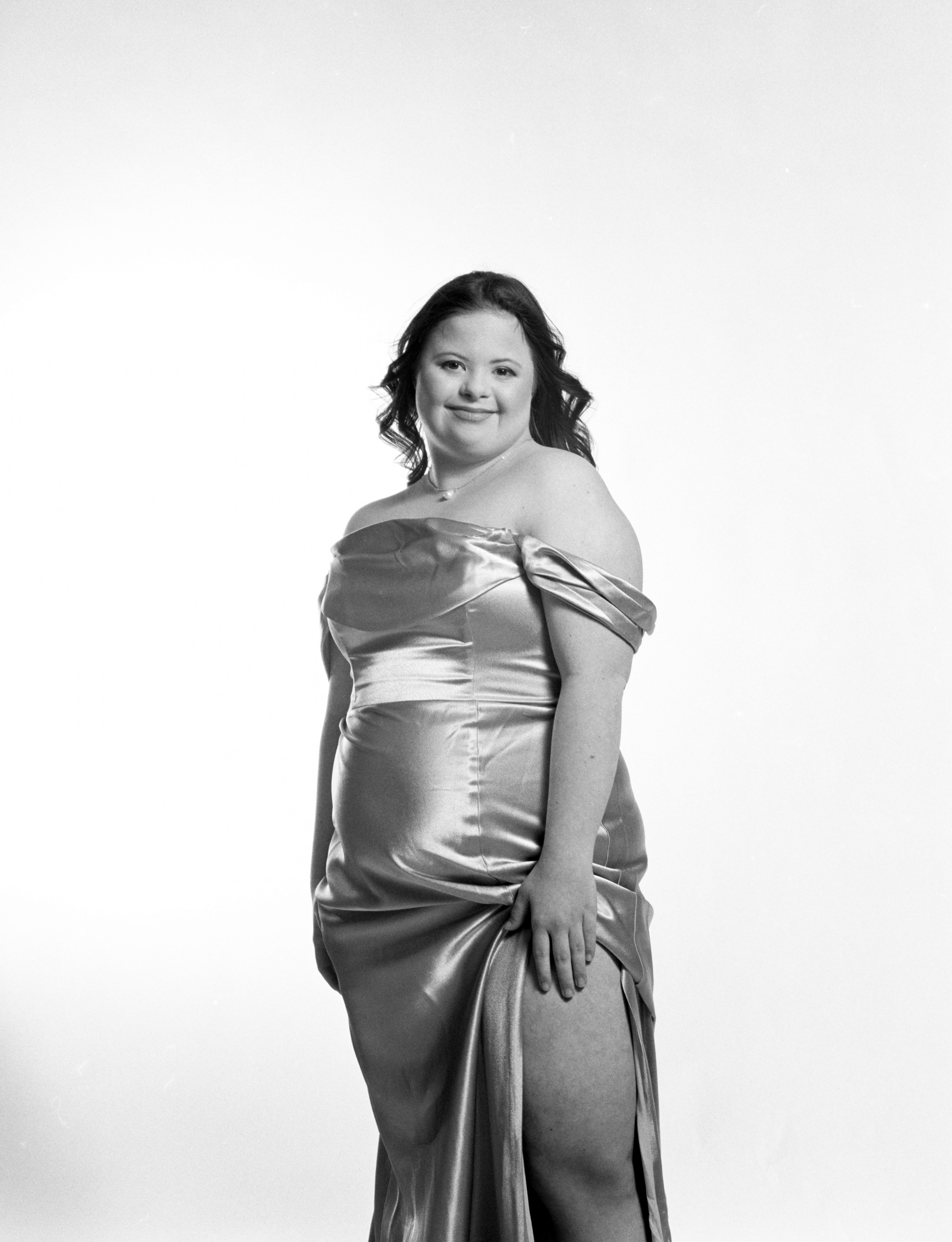 Dramatic black and white photo. Rachel, a short, white woman with down syndrome, poses softly while standing with her hands near her body in a strapless satin bridesmaids dress.