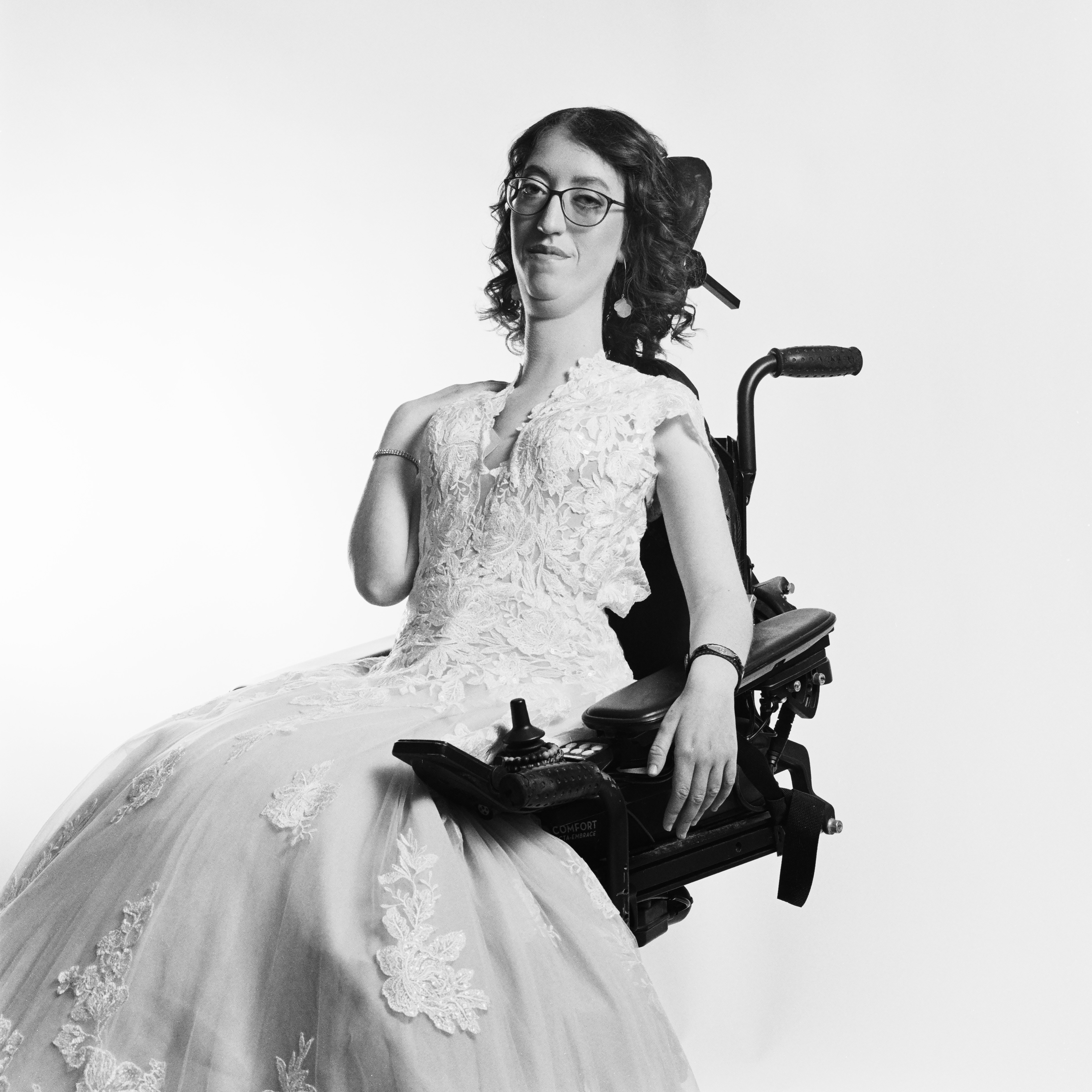 Dramatic black and white photo. Naomi, a young white woman with short curly hair wearing glasses, poses with one hand on her shoulder and the other on her power wheelchair in a wedding dress.