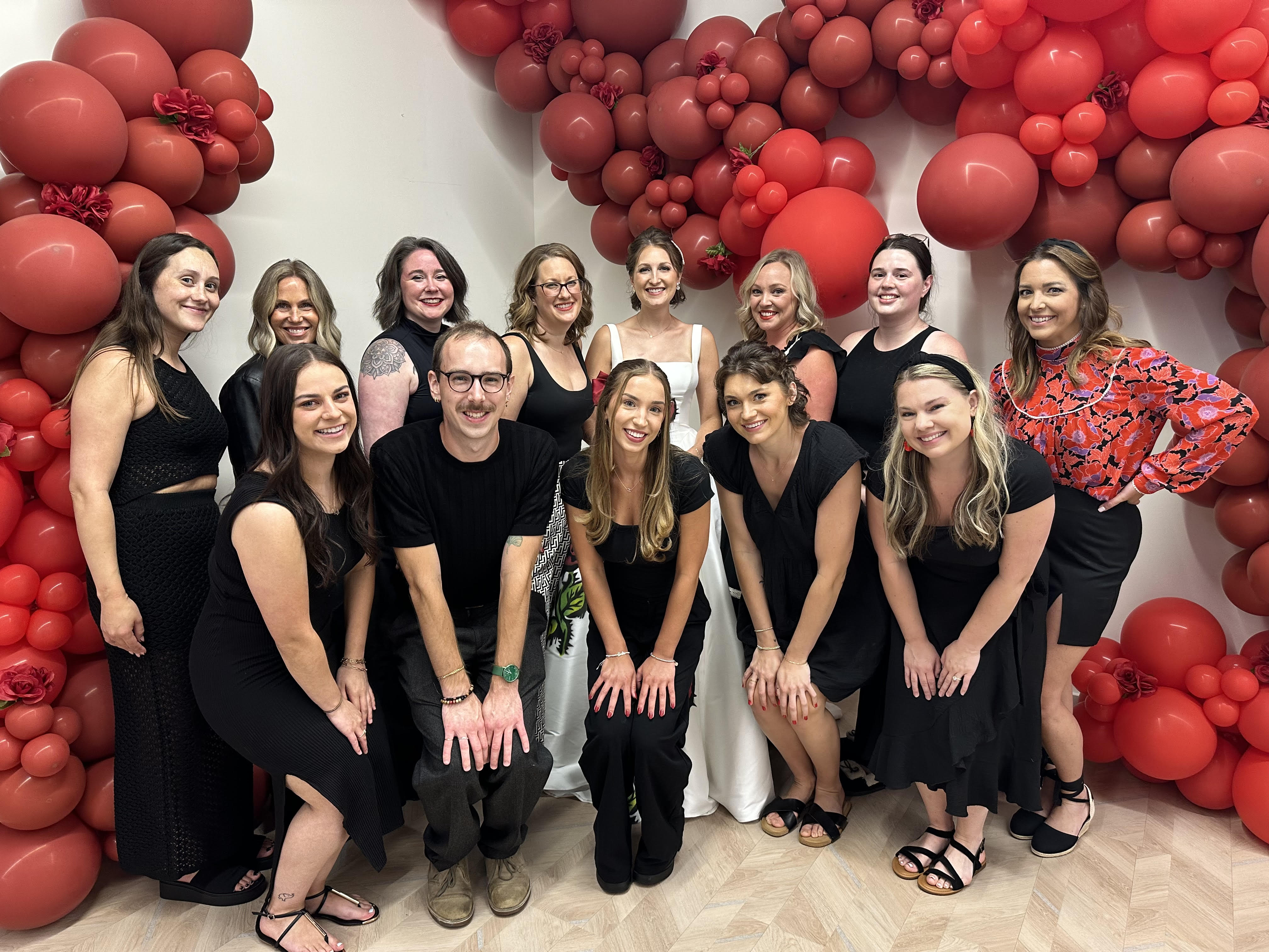 13 members of the Heart to Heart Bridal team pose in front of a balloon wall.