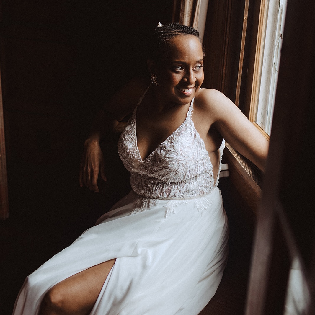 Danica, a black woman with her hair pulled back in a updo, sitting by a windowsill in a wedding dress. Photo has moody lighting.